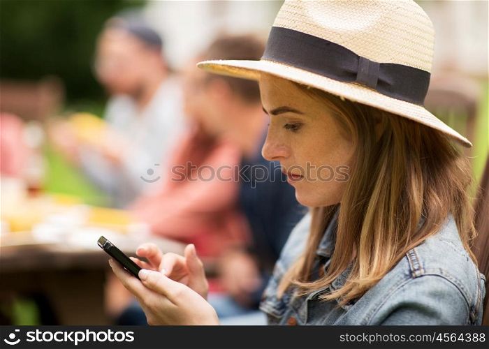 leisure, holidays, people and technology concept - close up of young woman or teenage girl texting on smartphone and friends having dinner at summer garden party