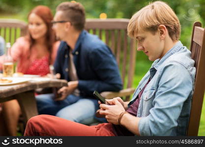 leisure, holidays, people and technology concept - young man texting on smartphone and friends having dinner at summer garden party