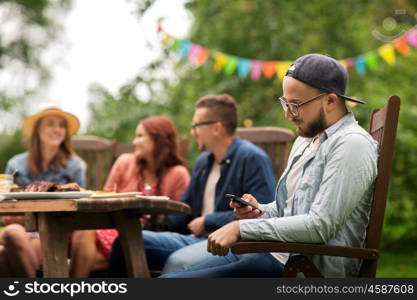 leisure, holidays, people and technology concept - young man texting on smartphone and friends having dinner at summer garden party