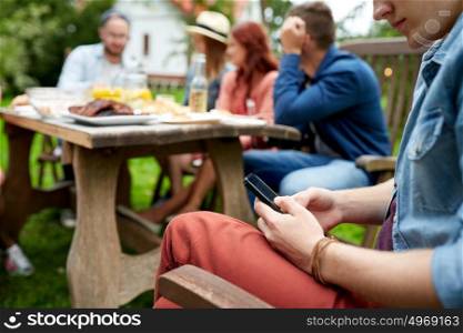 leisure, holidays, people and technology concept - young man texting on smartphone and friends having dinner at summer garden party. man with smartphone and friends at summer party