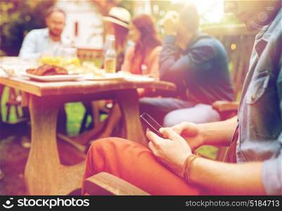 leisure, holidays, people and technology concept - young man texting on smartphone and friends having dinner at summer garden party. man with smartphone and friends at summer party