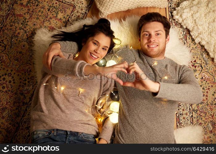 leisure, hygge and people concept - happy couple with garland lying on floor at home and making hand heart gesture. happy couple lying on floor and making hand heart