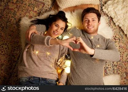 leisure, hygge and people concept - happy couple with garland lying on floor at home and making hand heart gesture. happy couple lying on floor and making hand heart
