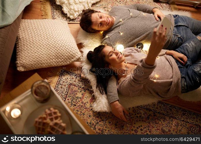 leisure, hygge and people concept - happy couple with garland lying on floor at home and making selfie by smartphone. couple lying on floor and making selfie at home