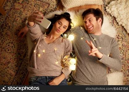 leisure, hygge and people concept - happy couple with garland lying on floor at home and making selfie by smartphone. couple lying on floor and making selfie at home