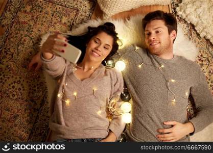 leisure, hygge and people concept - happy couple with garland lying on floor at home and making selfie by smartphone. couple lying on floor and making selfie at home. couple lying on floor and making selfie at home