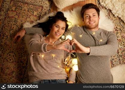 leisure, hygge and people concept - happy couple with garland lying on floor at home and making hand heart gesture. happy couple lying on floor and making hand heart. happy couple lying on floor and making hand heart