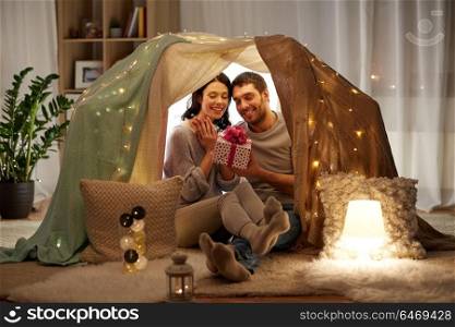 leisure, hygge and people concept - happy couple with gift box in kids tent at home. happy couple with gift box in kids tent at home
