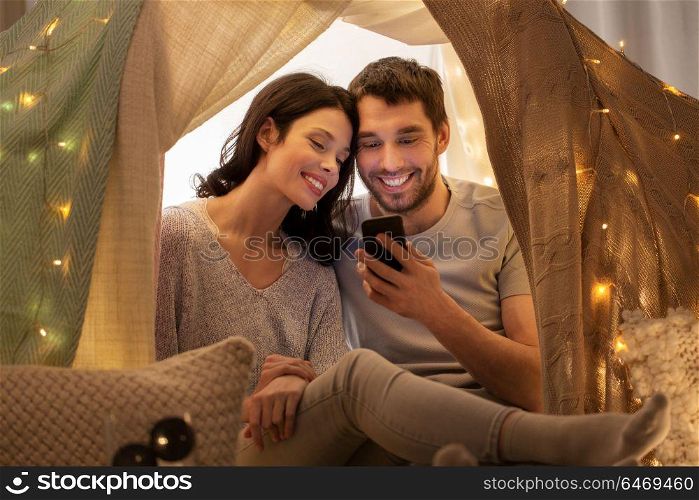leisure, hygge, technology and people concept - happy couple with smartphone in kids tent at home. happy couple with smartphone in kids tent at home