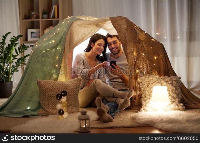 leisure, hygge, technology and people concept - happy couple with smartphone in kids tent at home. happy couple with smartphone in kids tent at home