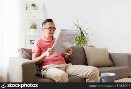 leisure, information, people and mass media concept - happy asian man in glasses reading newspaper at home. asian man in glasses reading newspaper at home