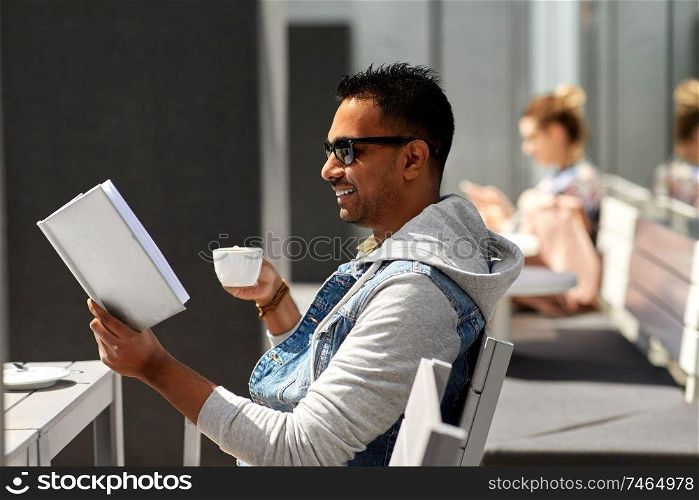 leisure, literature and people concept - indian man reading book and drinking takeaway coffee at street city cafe. man reading book and drinking coffee at city cafe
