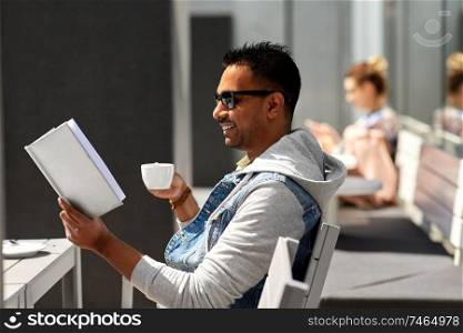 leisure, literature and people concept - indian man reading book and drinking takeaway coffee at street city cafe. man reading book and drinking coffee at city cafe