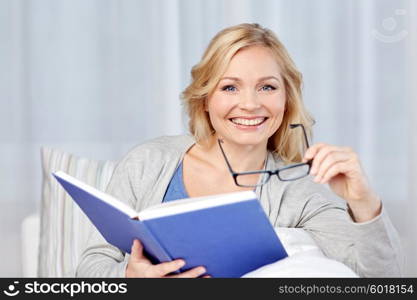 leisure, literature and people concept - smiling middle aged woman reading book and sitting on couch at home