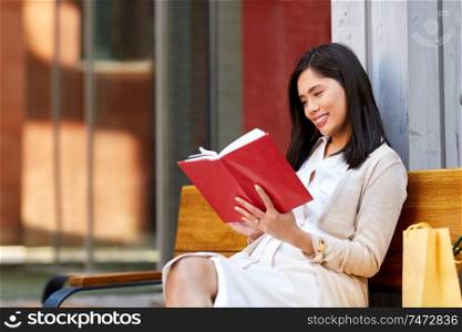 leisure, literature and people concept - smiling young asian woman reading book sitting on bench in city yard. smiling asian woman reading book sitting on bench