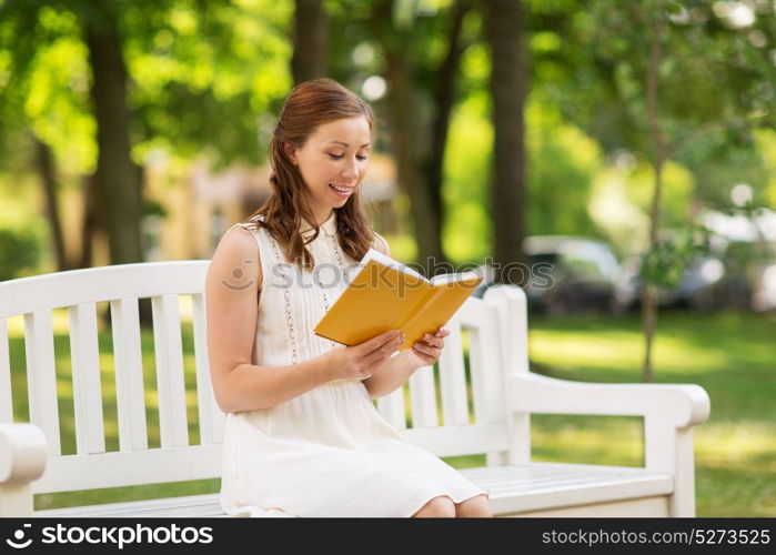 leisure, literature and people concept - smiling young woman in white dress reading book sitting on bench at summer park. smiling young woman reading book at summer park