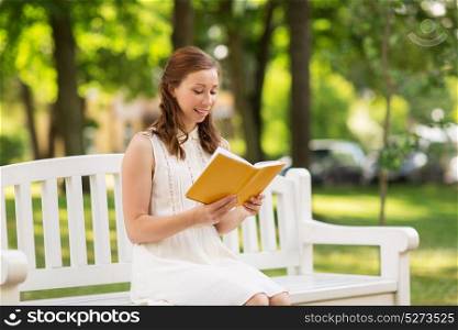 leisure, literature and people concept - smiling young woman in white dress reading book sitting on bench at summer park. smiling young woman reading book at summer park