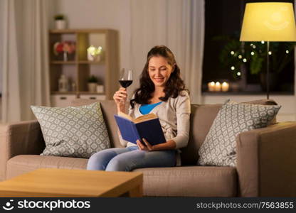 leisure, literature and people concept - young woman reading book and drinking red wine at home in evening. young woman reading book and drinking wine at home