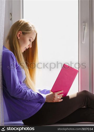 Leisure, literature and people concept. Young woman teen girl reading book at home while sitting on window sill. Calm and coziness.. Woman sitting on window sill reading book at home