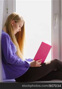Leisure, literature and people concept. Young woman teen girl reading book at home while sitting on window sill. Calm and coziness.. Woman sitting on window sill reading book at home