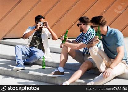 leisure, male friendship and people concept - man with camera photographing his friends drinking beer on street in summer. man photographing friends drinking beer on street