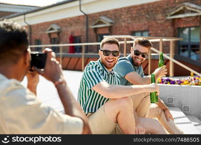 leisure, male friendship and people concept - man with camera photographing his friends drinking beer on street in summer. man photographing friends drinking beer on street