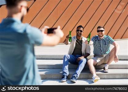 leisure, male friendship and people concept - man with smartphone photographing his friends toasting beer on street in summer. man photographing friends drinking beer on street