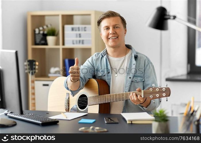 leisure, music and people concept - happy smiling young man or musician playing guitar sitting at table at home and showing thumbs up. happy man with guitar showing thumbs up at home