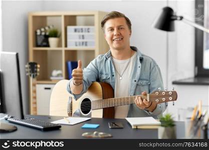leisure, music and people concept - happy smiling young man or musician playing guitar sitting at table at home and showing thumbs up. happy man with guitar showing thumbs up at home