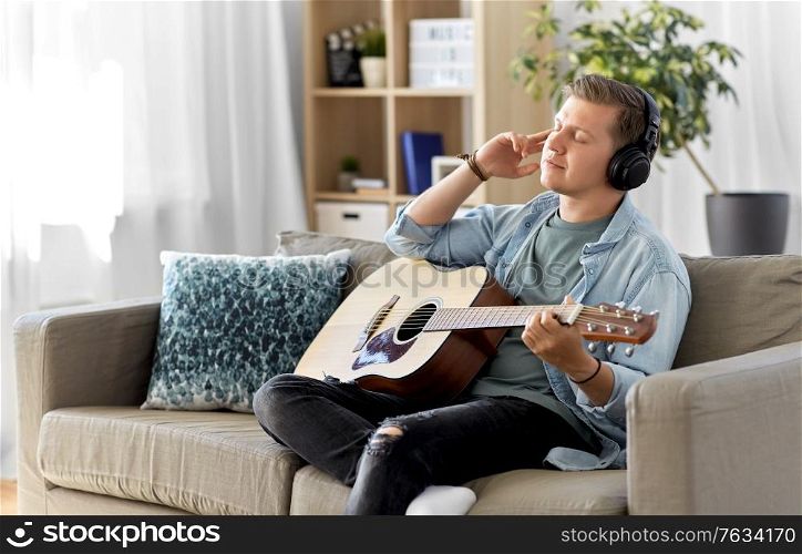 leisure, music and people concept - young man or musician in headphones playing guitar sitting on sofa at home. man in headphones playing guitar at home