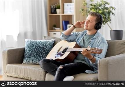 leisure, music and people concept - young man or musician in headphones playing guitar sitting on sofa at home. man in headphones playing guitar at home
