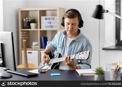leisure, music and people concept - young man or musician in headphones with computer playing bass guitar sitting at table at home. man in headphones playing bass guitar at home