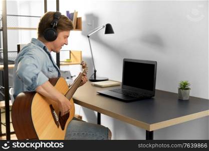 leisure, music and people concept - young man or musician in headphones with laptop computer playing guitar sitting at table at home. man in headphones playing guitar at home