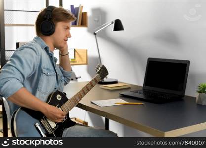 leisure, music and people concept - young man or musician in headphones with laptop computer playing bass guitar sitting at table at home. man in headphones playing bass guitar at home