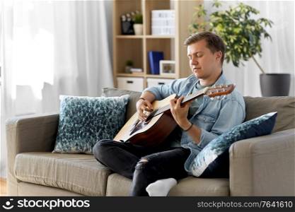 leisure, music and people concept - young man or musician playing guitar sitting on sofa at home. young man playing guitar sitting on sofa at home