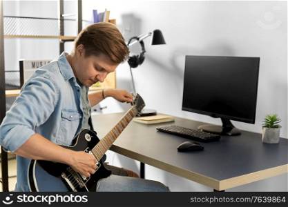 leisure, music and people concept - young man or musician with computer playing bass guitar sitting at table at home. young man with computer playing guitar at home