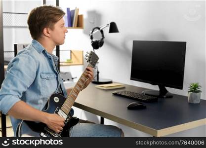 leisure, music and people concept - young man or musician with computer playing bass guitar sitting at table at home. young man with computer playing guitar at home