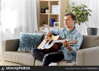 leisure, music and people concept - young man or musician with guitar, earphones and smartphone sitting on sofa at home. man with guitar, earphones and smartphone at home