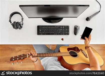 leisure, music and people concept - young man or musician with guitar, smartphone and computer sitting at table. young man with guitar and smartphone at table