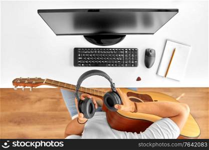 leisure, music and people concept - young man or musician with headphones, computer and guitar sitting at table. young man with headphones and guitar at table