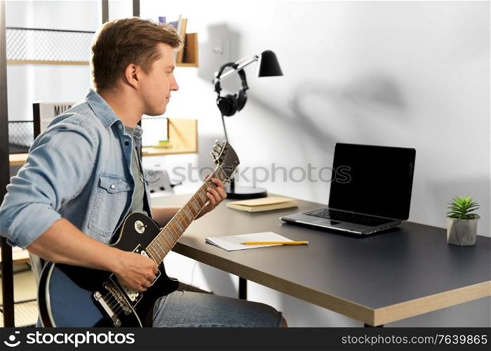 leisure, music and people concept - young man or musician with laptop computer playing bass guitar sitting at table at home. young man with laptop playing guitar at home