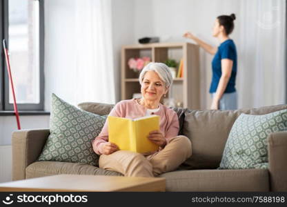 leisure, old age and people concept - happy senior woman reading book and housekeeper cleaning at home. old woman reading book and housekeeper at home