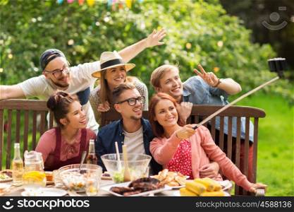 leisure, party, technology, people and holidays concept - happy friends taking picture with smartphone selfie stick and gathering for dinner at summer garden party