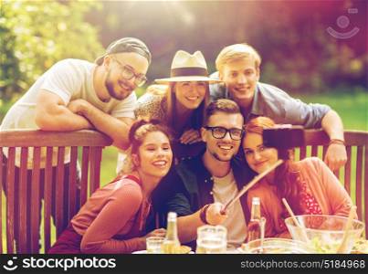 leisure, party, technology, people and holidays concept - happy friends taking picture with smartphone selfie stick and gathering for dinner at summer garden party. friends taking selfie at party in summer garden