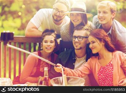 leisure, party, technology, people and holidays concept - happy friends taking picture with smartphone selfie stick and gathering for dinner at summer garden party. friends taking selfie at party in summer garden