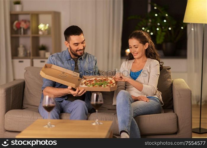 leisure, people and fast food concept - happy couple with wine eating takeaway pizza at home in evening. happy couple eating takeaway pizza at home