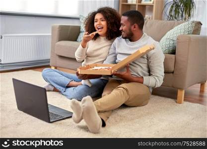 leisure, people and food concept - happy african american couple with laptop computer eating takeaway pizza at home. happy african american couple eating pizza at home