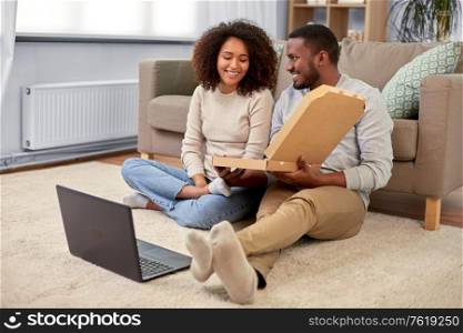 leisure, people and food concept - happy african american couple with laptop computer eating takeaway pizza at home. happy african american couple eating pizza at home
