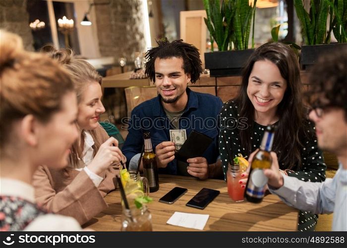 leisure, people and holidays concept - happy friends with drinks, money and bill at bar or cafe