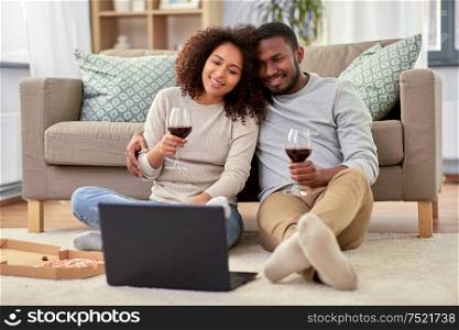 leisure, people and technology concept - happy african american couple with laptop computer and takeaway pizza and drinking red wine at home. happy couple with laptop drinking red wine at home